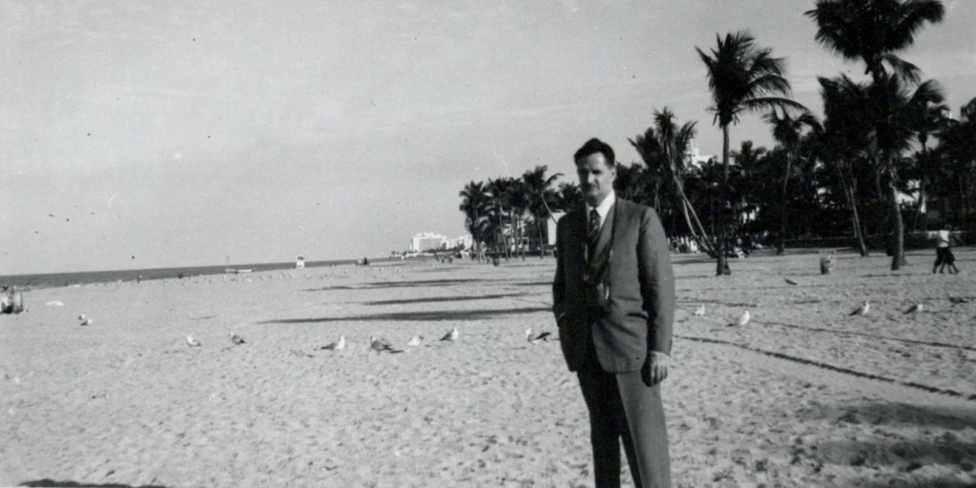 Black and white archival photo of John Blume, standing on a beach in a suit looking towards the camera.